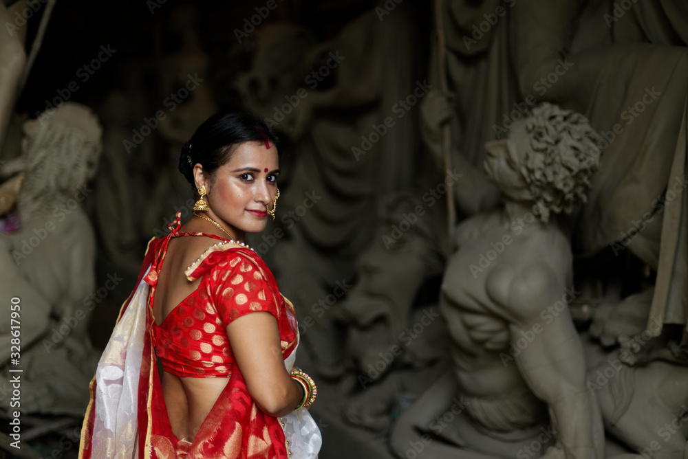 An young and beautiful Indian Bengali brunette woman in red and white traditional ethnic sari worshiping clay idol of Hindu goddess Durga while turning back. Indian culture, religion and fashion