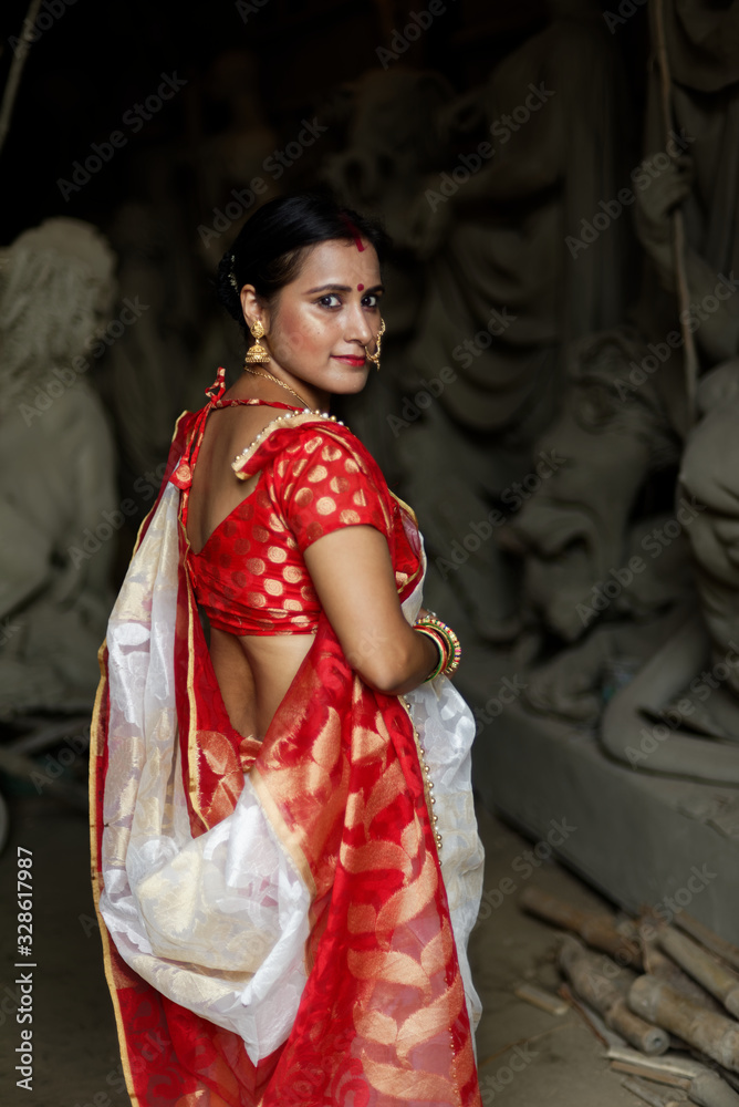 An young and beautiful Indian Bengali brunette woman in red and white traditional ethnic sari worshiping clay idol of Hindu goddess Durga while turning back. Indian culture, religion and fashion