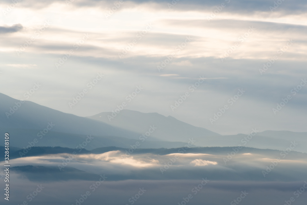 clouds and rays of dawn in mountain valley, soft light in early morning, meditation in nature. Gentle hills in bluish haze, silhouettes of mountains