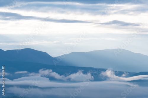 clouds and rays of dawn in mountain valley, soft light in early morning, meditation in nature. Gentle hills in bluish haze, silhouettes of mountains © Koirill