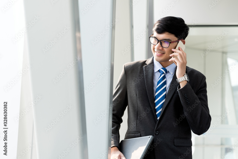 A asian handsome young businessman holding his laptop tablet while using his phone and standing on the escalator. Business concept. A asian handsome young businessman holding his laptop tablet while using his phone and standing on the escalator. Business concept.