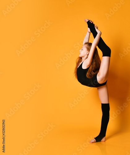 Smiling teen girl rhythmic gymnast does vertical splits holding her leg up with both hands over her head looking forward