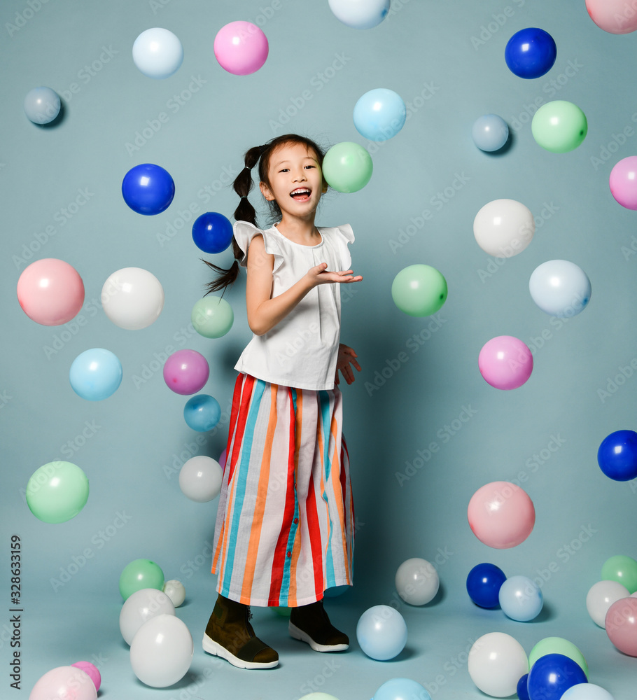 Fototapeta premium Laughing asian kid girl in party clothes is having fun surrounded by colorful air balloons holds hand up with open palm