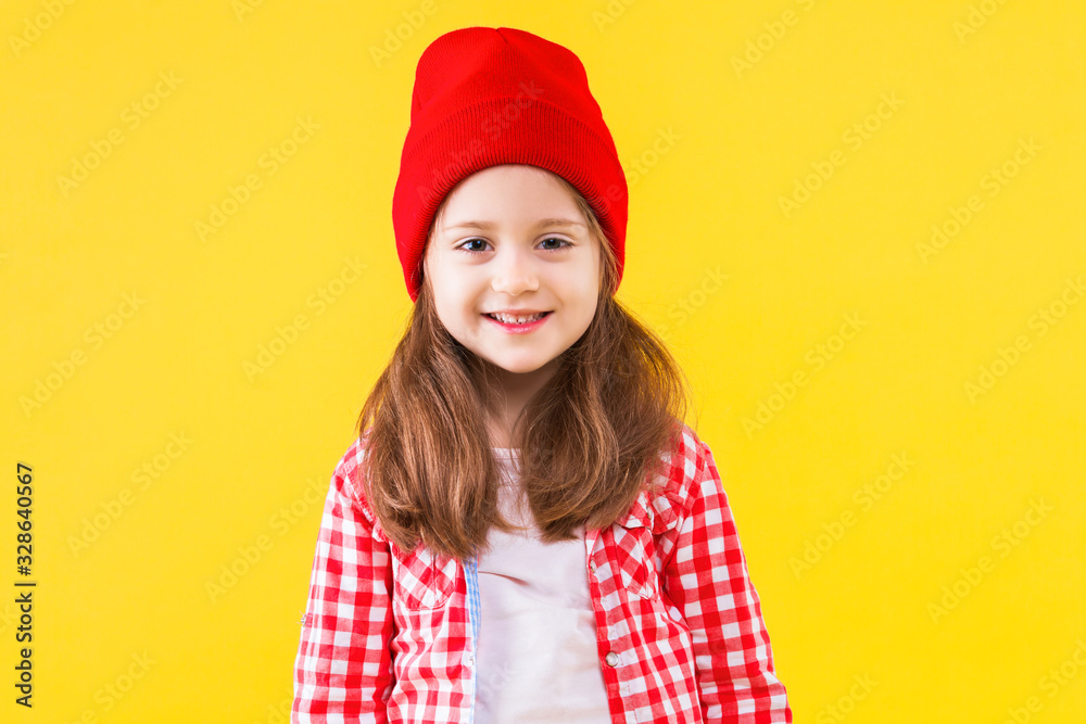 Cheerful stylish pretty little school girl dressed in pink checkered shirt, red cap, hat on yellow orange background. Cute child pupil is smiling and having fun. Emotional portrait concept. Cheerful stylish pretty little school girl dressed in pink checkered shirt, red cap, hat on yellow orange background. Cute child pupil is smiling and having fun. Emotional portrait concept.