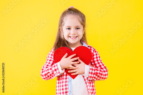 Pretty fair haired girl in pink checkered shirt is holding in hand red paper heart sign. Cute child is smiling on yellow orange background. Emotional portrait concept. Saint Valentine day.