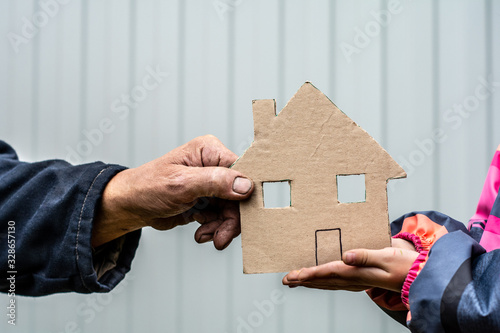 A man's hand holds a paper house cut out of cardboard. Nearby, children's hands hold the house from below.