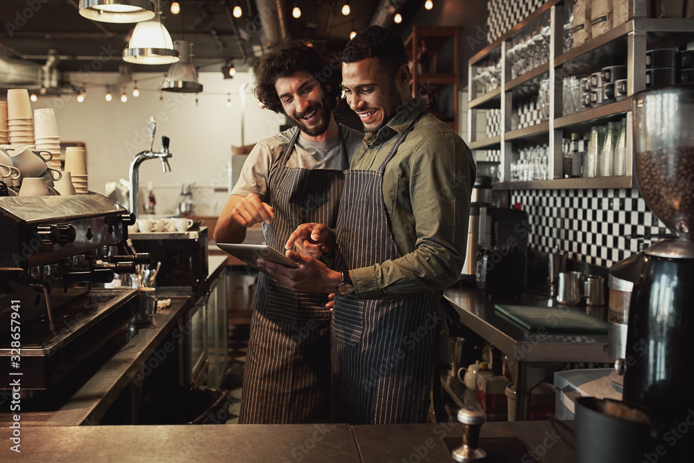 Smiling handsome waiter holding tablet with coworker while pointing on it in coffee shop sharing recipe