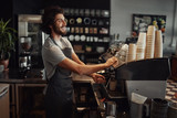 Young cheerful barista wearing black apron while preparing coffee at an automatic machine in a modern coffee shop