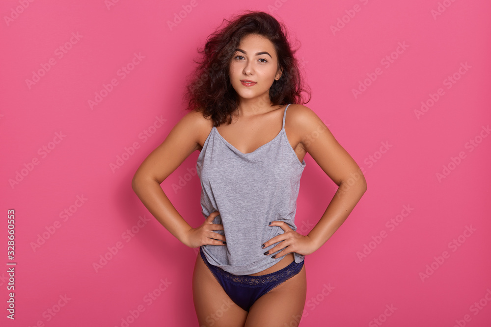 Beautiful woman wearing blue underwear and gray cotton t shirt standing isolated over rose studio background, having dark wavy hair, female with prfect body keeping hands on hips, looking at camera. Beautiful woman wearing blue underwear and gray cotton t shirt standing isolated over rose studio background, having dark wavy hair, female with prfect body keeping hands on hips, looking at camera.