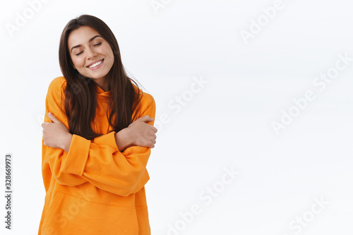 Tenderness and women concept. Adorable caucasian female brunette in orange hoodie, close eyes and smiling lovely, embrace herself, cuddle feeling cozyness soft hoodie, white background photo