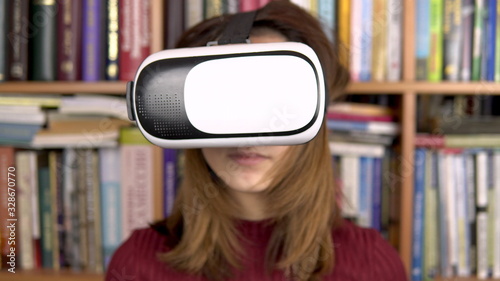 A young woman reads a book with BP glasses in the library. A woman with a VR helmet on her head. In the background are books on bookshelves. Book library.