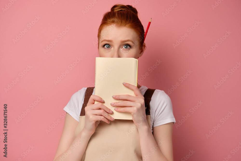 A red-haired girl in an apron with collected hair stands on a pink background, covering her mouth with a notebook and wide-eyed A red-haired girl in an apron with collected hair stands on a pink background, covering her mouth with a notebook and wide-eyed