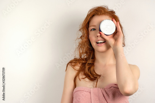 A red-haired curly-haired girl in a pink towel stands on a white background, covering her eye with a jar of cream