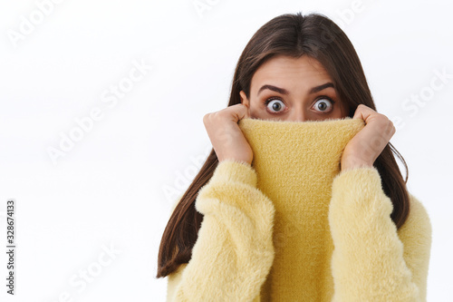 Close-up portrait of surprised startled cute girl hiding face in sweater collar, pulling it on nose and express shock with eyes, staring speechless camera afraid or scared, white background