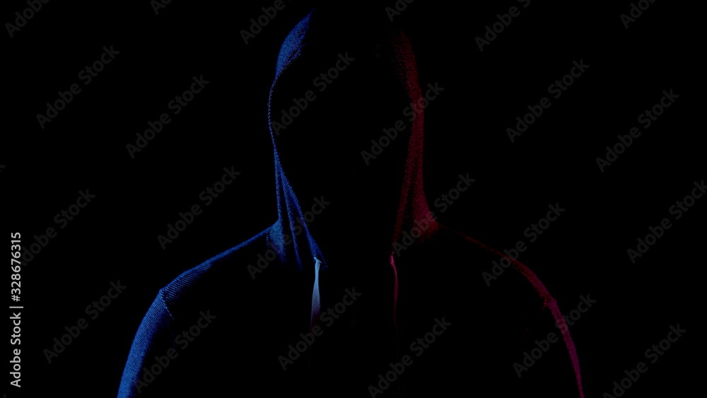 A young man is standing in the hood hiding his face. An attacker is standing in the dark. Blue and red light falls on the person on a black background. A young man is standing in the hood hiding his face. An attacker is standing in the dark. Blue and red light falls on the person on a black background.