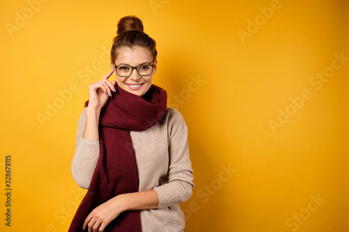 A beautiful brunette with a bun on head stands in a sweater and a burgundy scarf on a yellow background, straightens glasses