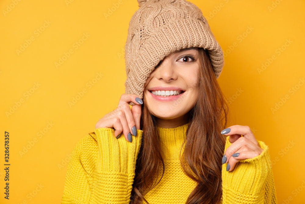 A brunette in a yellow sweater is standing on a yellow background, smiling and pulling her hat over half of her face. A brunette in a yellow sweater is standing on a yellow background, smiling and pulling her hat over half of her face.