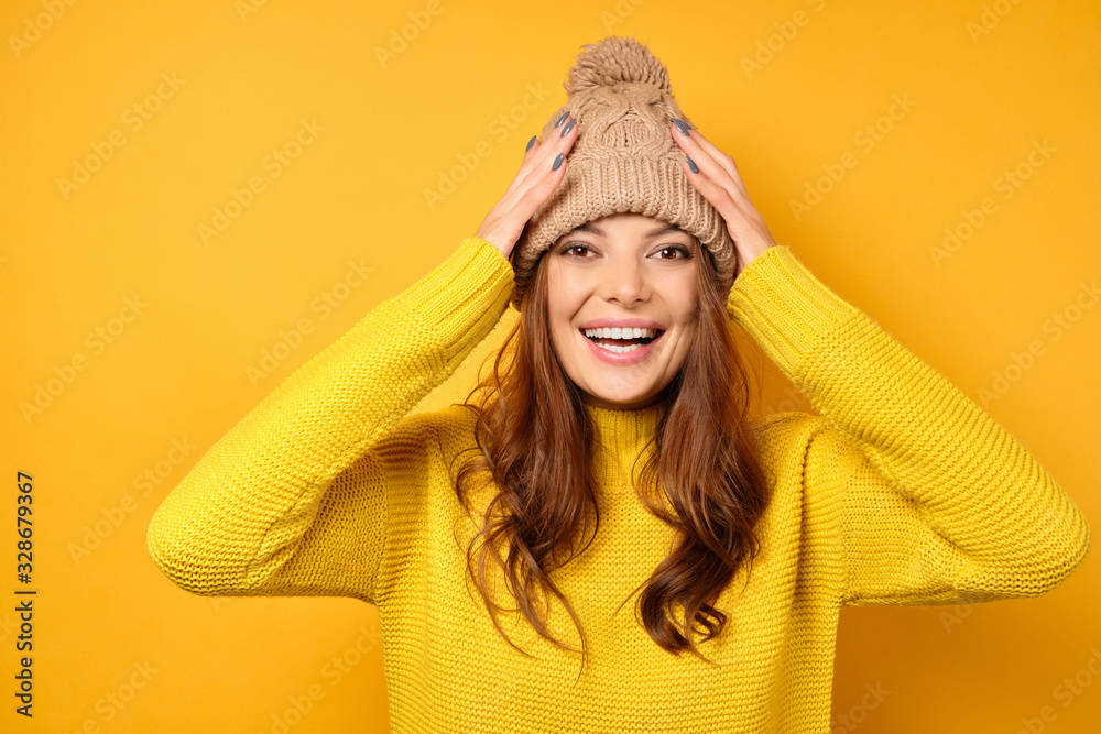A brunette in a yellow sweater stands on a yellow background and laughs, adjusting her hat. A brunette in a yellow sweater stands on a yellow background and laughs, adjusting her hat.