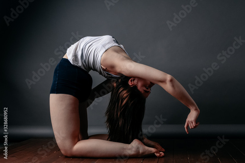 Beautiful woman standing in camel pose on a wooden floor doing Ustrasana exercises, practicing yoga, a sports girl in black sports shorts and a top, exercising home in yoga studio with gray background