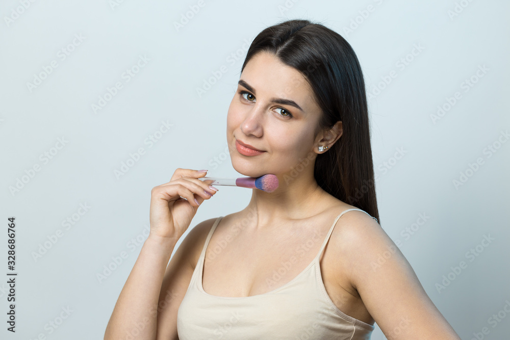 Close-up of a young girl in a light top on a white background making a facial make-up. A pretty woman holds a cosmetic brush near her face and smiles. Close-up of a young girl in a light top on a white background making a facial make-up. A pretty woman holds a cosmetic brush near her face and smiles.