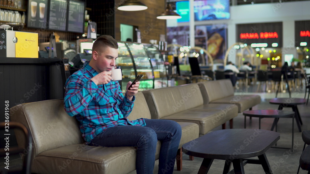 A young man is sitting in a cafe with a phone. The man is in correspondence on a smartphone and drinks coffee. Close-up. A young man is sitting in a cafe with a phone. The man is in correspondence on a smartphone and drinks coffee. Close-up.