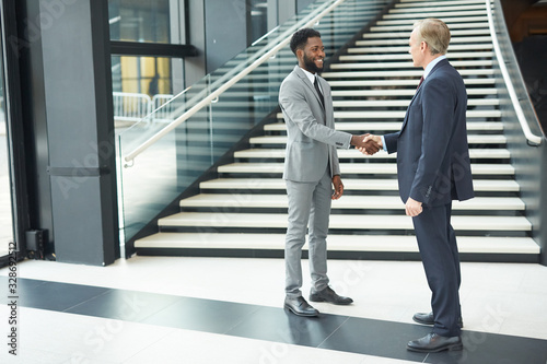Horizontal full shot of two cheerful modern businessmen standing in front of each other shaking their hands