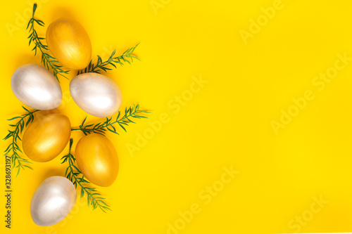 Eggs of silver and gold, green twigs from the left edge of the image on a plain yellow background. Easter holiday card. Copyspace.