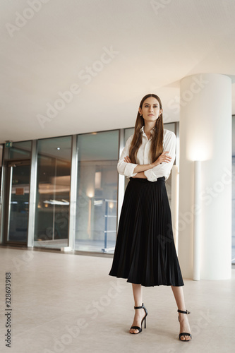 Vertical shot of proud and confident, successful businesswoman in company office, standing assured with crossed hands, looking pleased on distanse as thinking new deals and productive projects