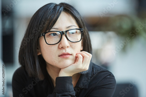 Cloe up head and shoulders portrait shot of attactive young Asian woman wearing stylish eyeglasses resting chin on her hand looking away photo