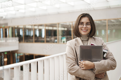 Education, career and university concept. Portrait of happy young pretty girl in jacket and glasses, holding laptop with worksheets and books, leaning on staircase and gazing away cheerful