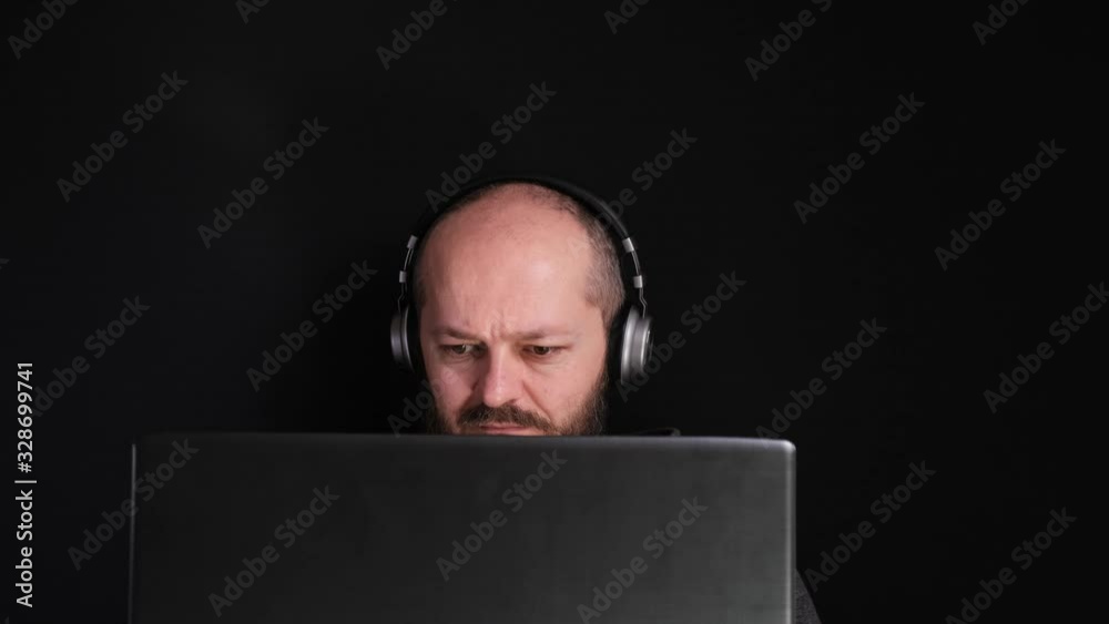 Working from home. Caucasian man in hoodie and headphones sitting behind the laptop and working at home, black background, home office concept Working from home. Caucasian man in hoodie and headphones sitting behind the laptop and working at home, black background, home office concept