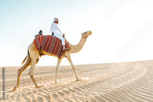 Photography Man wearing traditional clothes, taking a camel out on the desert sand, in Dubai