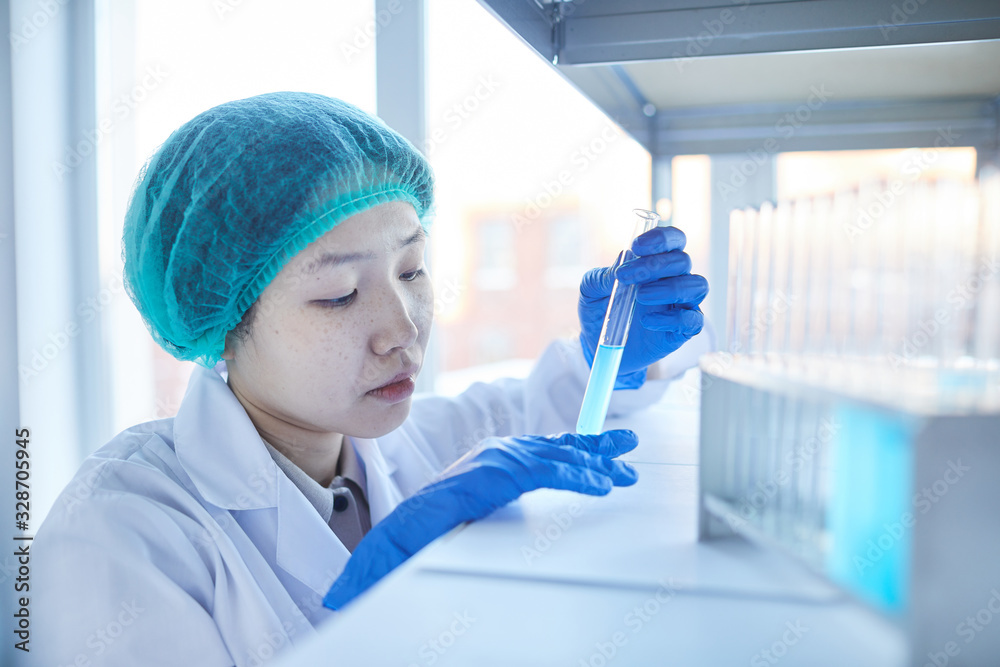 Horizontal close-up portrait of serious young Asian woman in lab coat watching chemical reaction in test-tube Horizontal close-up portrait of serious young Asian woman in lab coat watching chemical reaction in test-tube