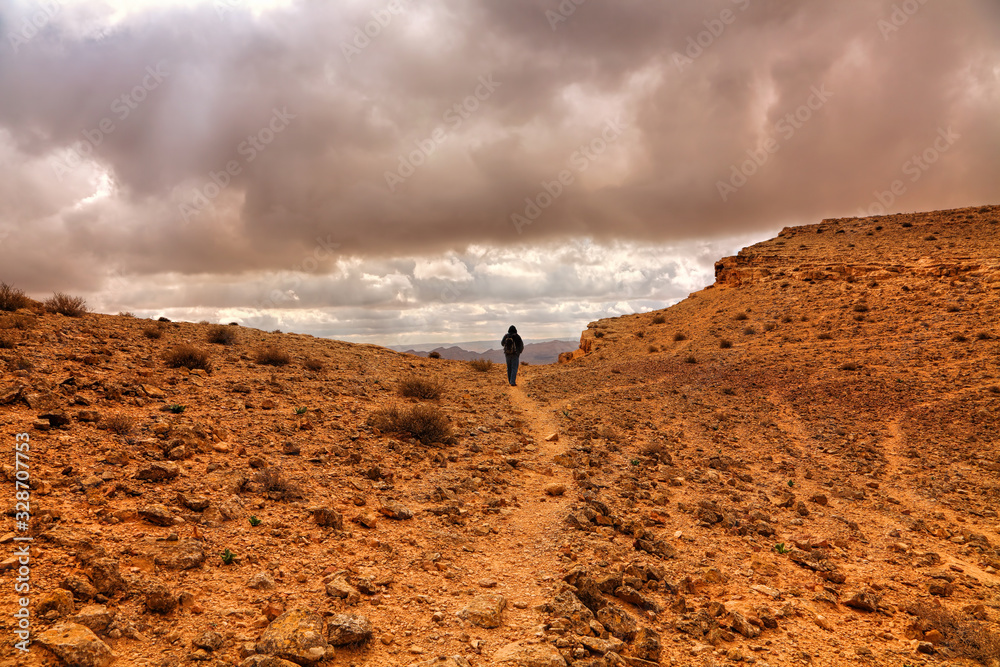 Lonely traveler hiking on rocky desert. Stormy heavy dark clouds above the ground. National geological park HaMakhtesh HaGadol. Big Crater, geological erosional land form. Negev desert. Israel Lonely traveler hiking on rocky desert. Stormy heavy dark clouds above the ground. National geological park HaMakhtesh HaGadol. Big Crater, geological erosional land form. Negev desert. Israel