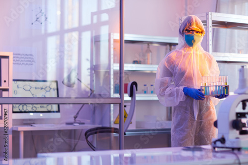 Unrecognizable female chemist wearing disposable protective uniform holding test-tubes with liquids looking at camera, horizontal medium long shot, copy space