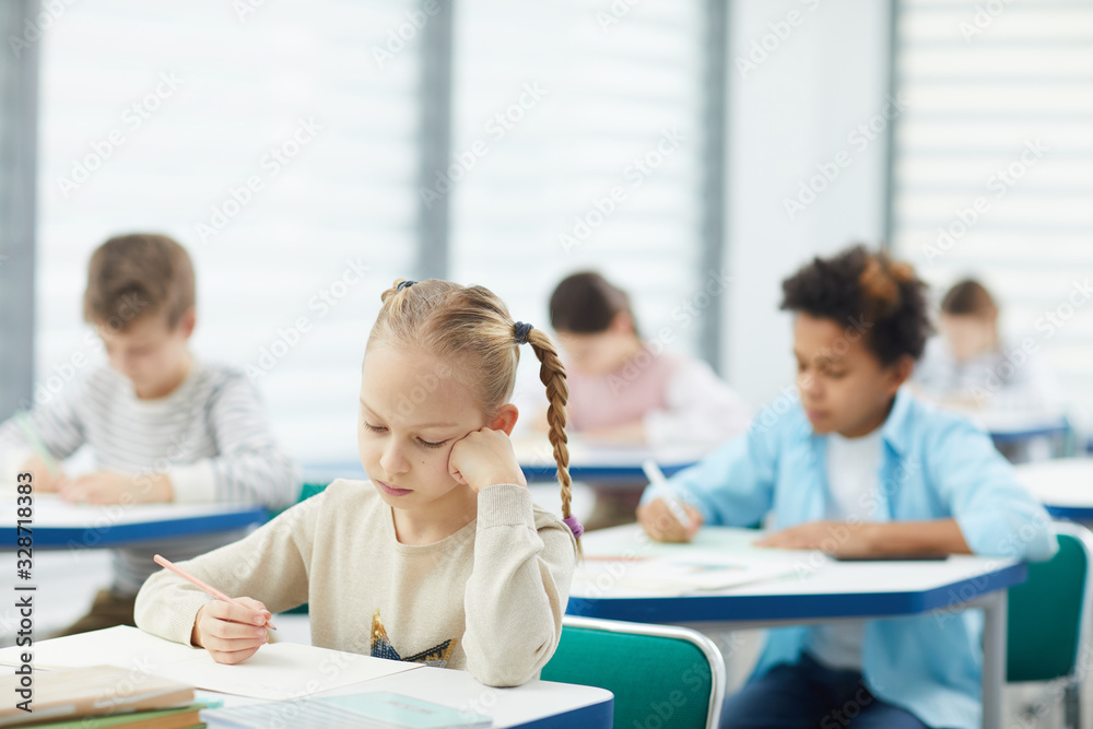 Horizontal portrait of little blond girl with two plaits sitting at school desk resting her cheek on hand learning boring lesson, copy space Horizontal portrait of little blond girl with two plaits sitting at school desk resting her cheek on hand learning boring lesson, copy space