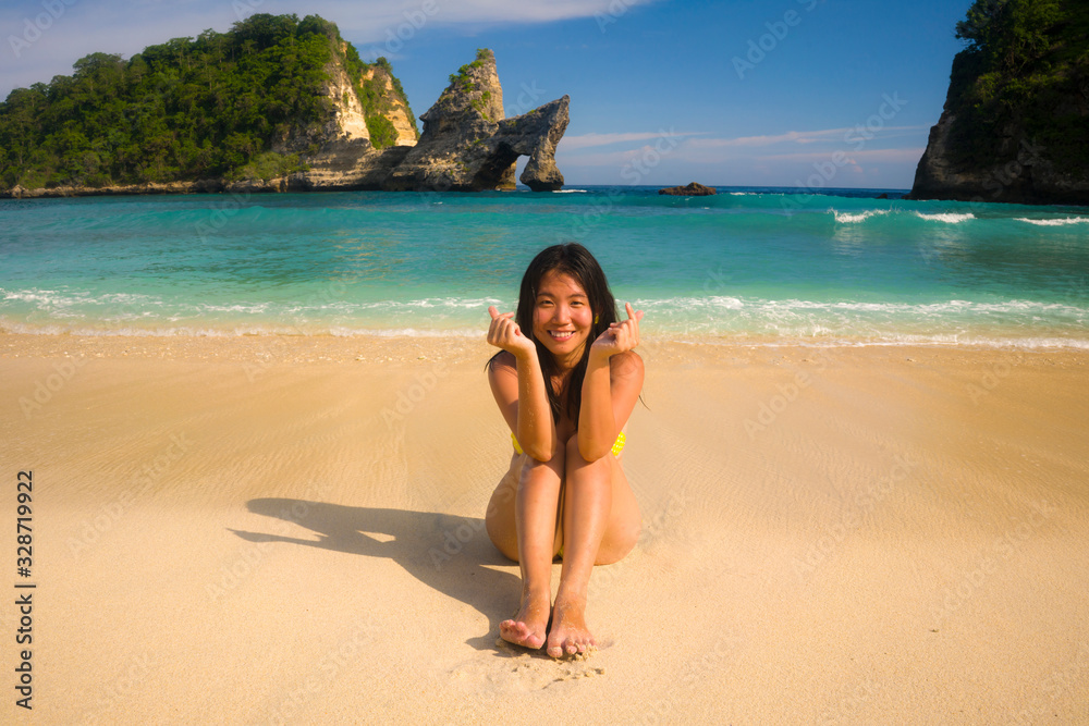 young happy and beautiful Asian Chinese woman in bikini excited having holidays at paradise beach island sitting on the sand by the sea enjoying idyllic travel destination