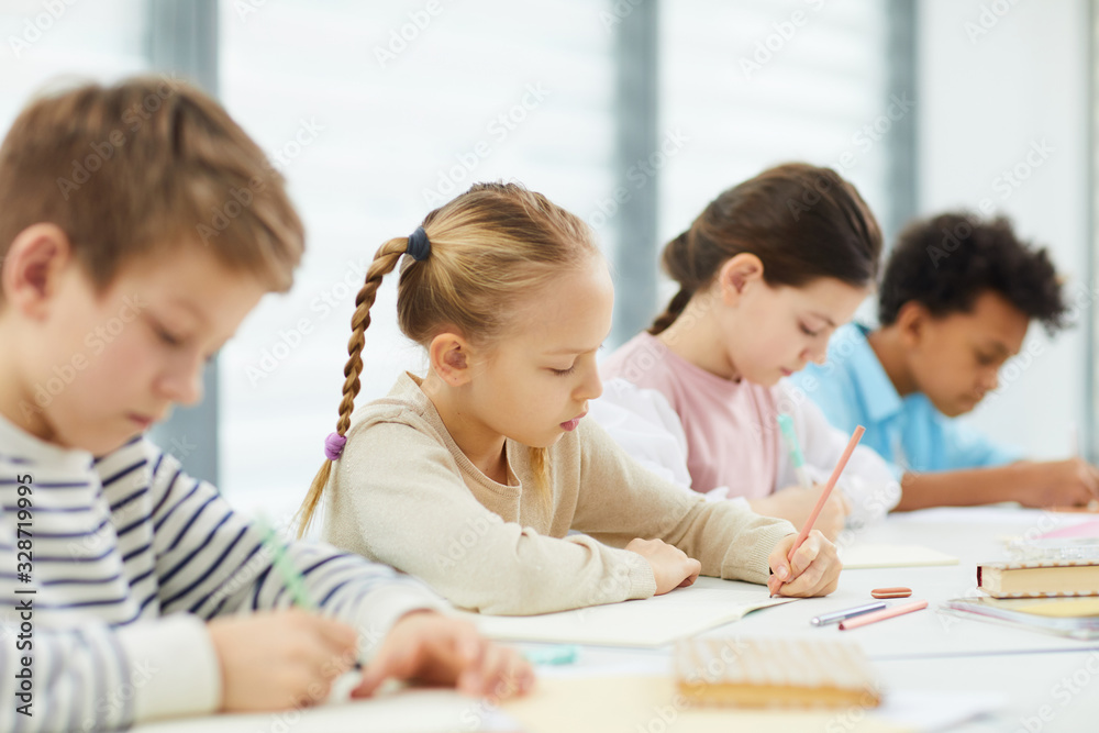 Horizontal portrait of four children wearing casual outfits sitting at desk in modern classroom writing something in their notebooks, copy space Horizontal portrait of four children wearing casual outfits sitting at desk in modern classroom writing something in their notebooks, copy space