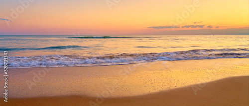 Colorful sunset at the tropical beach, sun behind clouds reflects on water and waves with foam hitting sand.