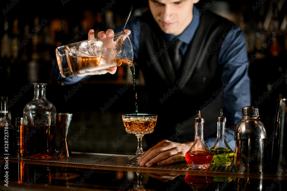 Young bartender pours a ready-made cold cocktail into wineglass.