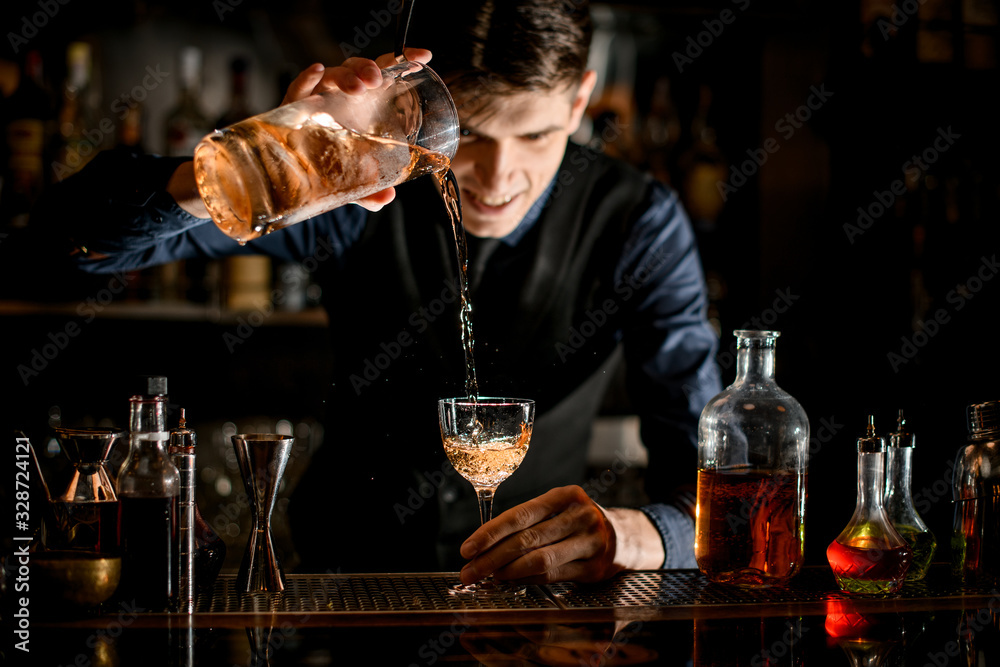 Bartender holds in one hand glass with a cocktail and pours it into wineglass in his other hand.