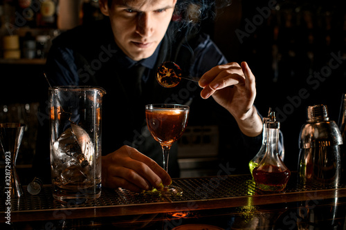 Young barman decorates glass with cocktail by burnt citrus slice.