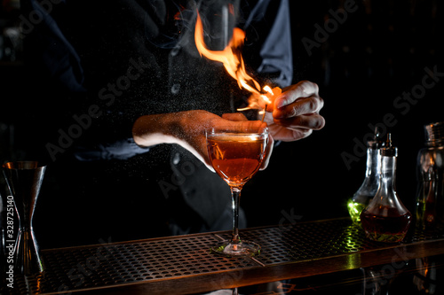 close-up of cocktail glass which bartender sprinkling by juice of citrus and sets fire.
