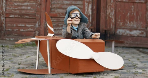 Cute joyful small Caucasian boy with red hair in hat and glasses sitting outdoor in wooden toy model of airplane and playing like rulling. Happy child dreaming to be aviator when grow up. photo
