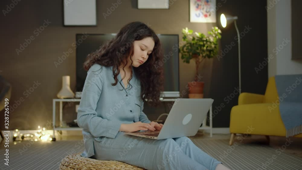 Calm cute young girl in light blue pajama using computer online at cozy home indoors, pretty curly pensive millennial female staying up to date in social media network chatting communicating
