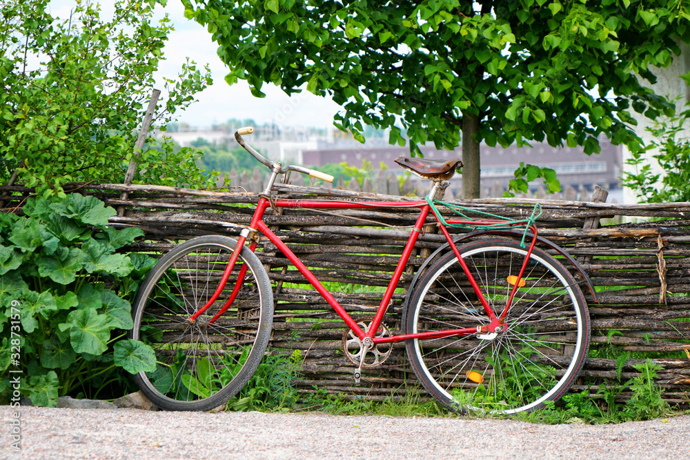 Vintage red bicycle parked near a lath wooden fence under a tree close to green bushes