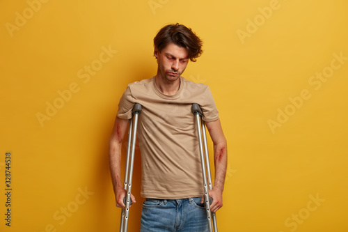 Sad unhappy man looks down, has serious injury after falling down from height, tired of long recovery period, tries to walk with crutches, poses against yellow background. Handicapped disabled male © wayhome.studio 
