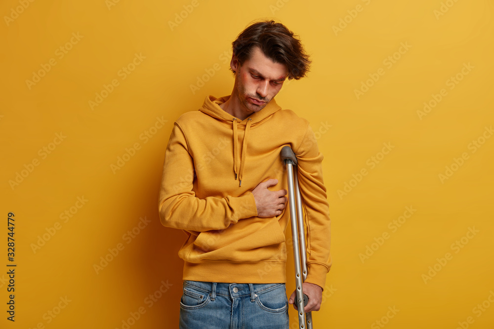 Indoor shot of distressed man has broken rib, suffers from pain, stands on crutches, had accident on road, wears yellow sweatshirt, has illness and injury, poses over yellow background. Mobility aid