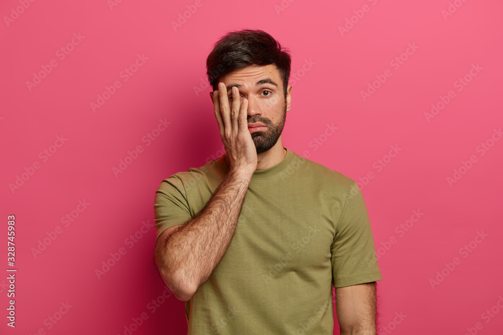 Tired bearded Caucasian man covers face with palm, looks with unhappy expression at camera, feels exhausted and sleepy, wears casual t shirt, poses over pink background, not eager to do something