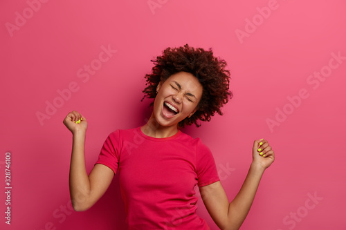 Photo of overjoyed woman triumphant makes fist bump, tilts head and laughs with joy, celebrates own success, wears casual t shirt, gets victory and achieves goal, poses over pink background.